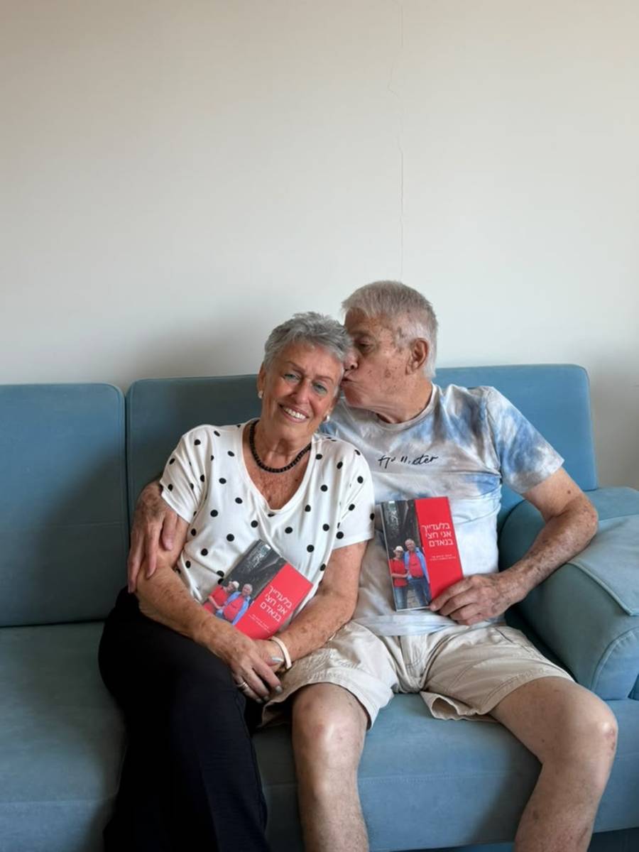 Nehemia kisses Adina's head on their sofa, each holding a copy of their printed couple's book.